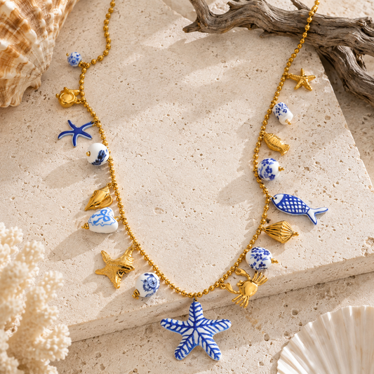 A gold-tone beaded necklace with nautical charms including blue and white ceramic seashells, starfish, fish, crabs, and a large blue-and-white starfish pendant, styled on a beige stone surface with driftwood and seashells.