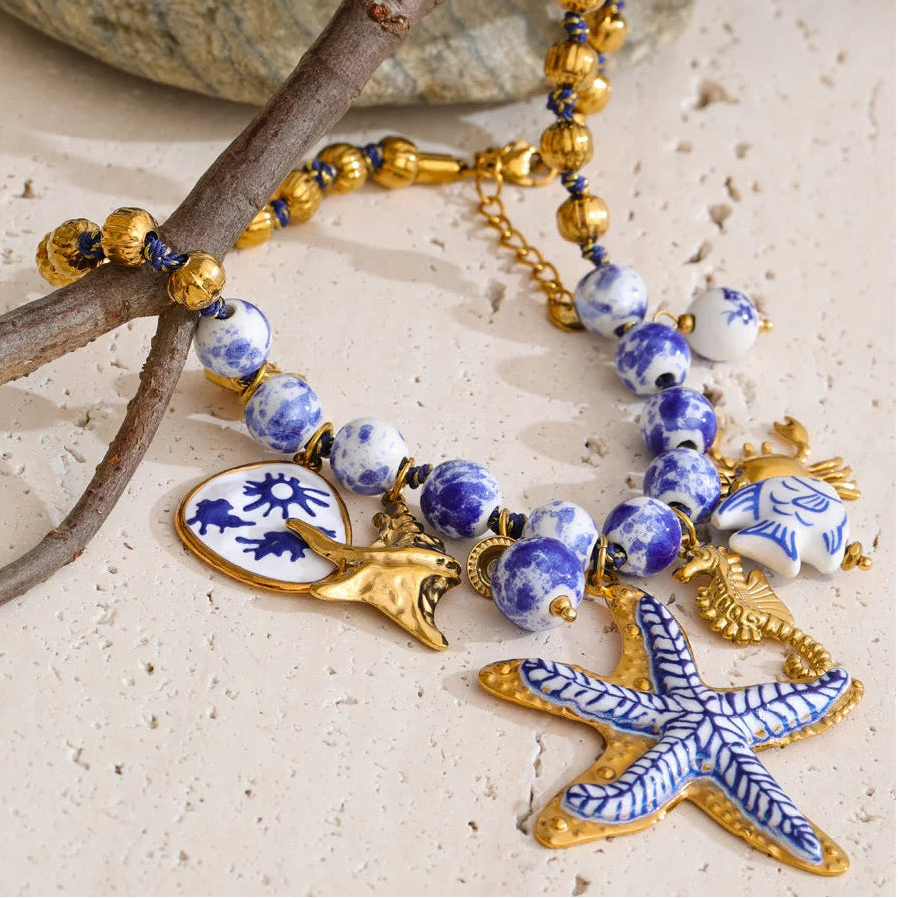 A decorative beaded necklace with blue and white marbled beads, gold-toned metal beads, and sea-themed charms including a starfish, seahorse, fish, and shell, displayed on a light stone surface with a branch prop.