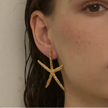 A close-up of a model wearing a textured gold starfish-shaped earring, paired with a light beige halter top against a neutral beige background.