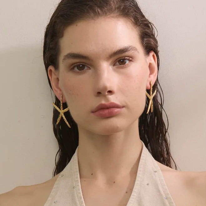A model with wet dark hair wearing a beige halter top with small rhinestone embellishments and gold star-shaped earrings, set against a plain beige background.