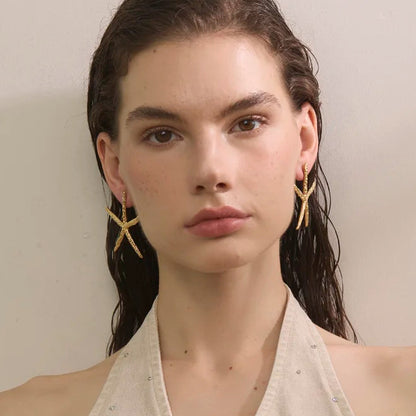 A model with wet dark hair wearing a beige halter top with small rhinestone embellishments and gold star-shaped earrings, set against a plain beige background.
