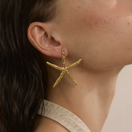 A close-up side profile of a woman wearing a textured gold starfish-shaped dangle earring against a soft beige background.