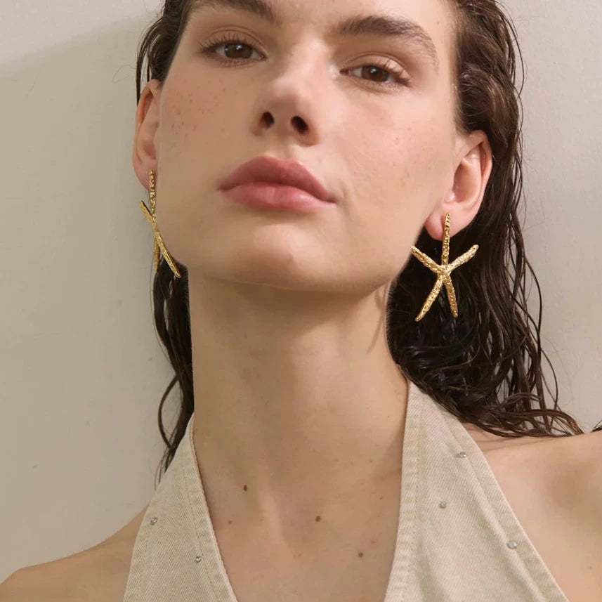 A close-up of a woman wearing gold starfish-shaped drop earrings and a light beige halter top against a plain light background.