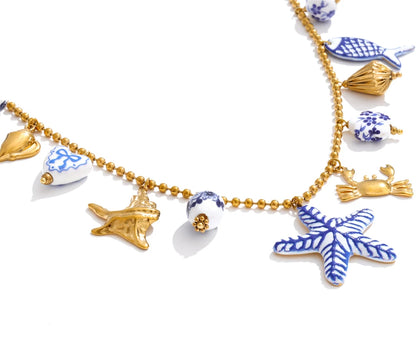 A gold beaded necklace featuring various ceramic nautical charms, including a blue and white starfish, crab, fish, and shell, displayed on a clean white background.